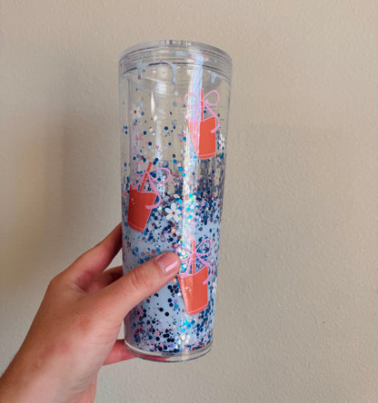 Clear tumbler with colorful glitter and iced coffee design held by a hand against a plain background